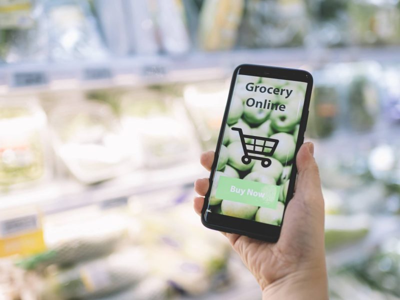 Knuspr: Eine Hand hält ein Handy, auf dem eine App für einen Online-Supermarkt geöffnet ist. Im Hintergrund ist unscharf ein Supermarktregal zu erkennen.