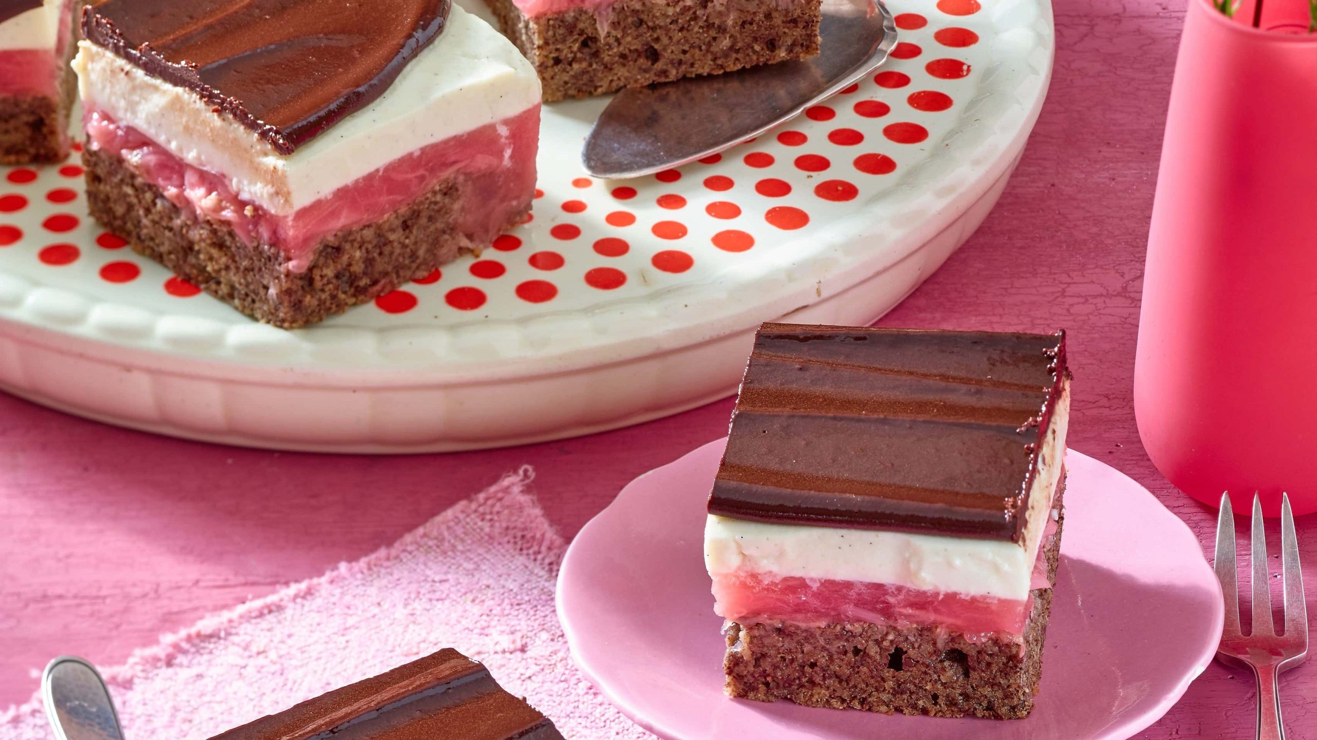Schokoladige Rhabarberschnitten mit weißer creme auf einem rosa Teller mit einer Gabel auf einem rosa Tisch neben einer Servierplatte mit weiteren Stücken Kuchen.