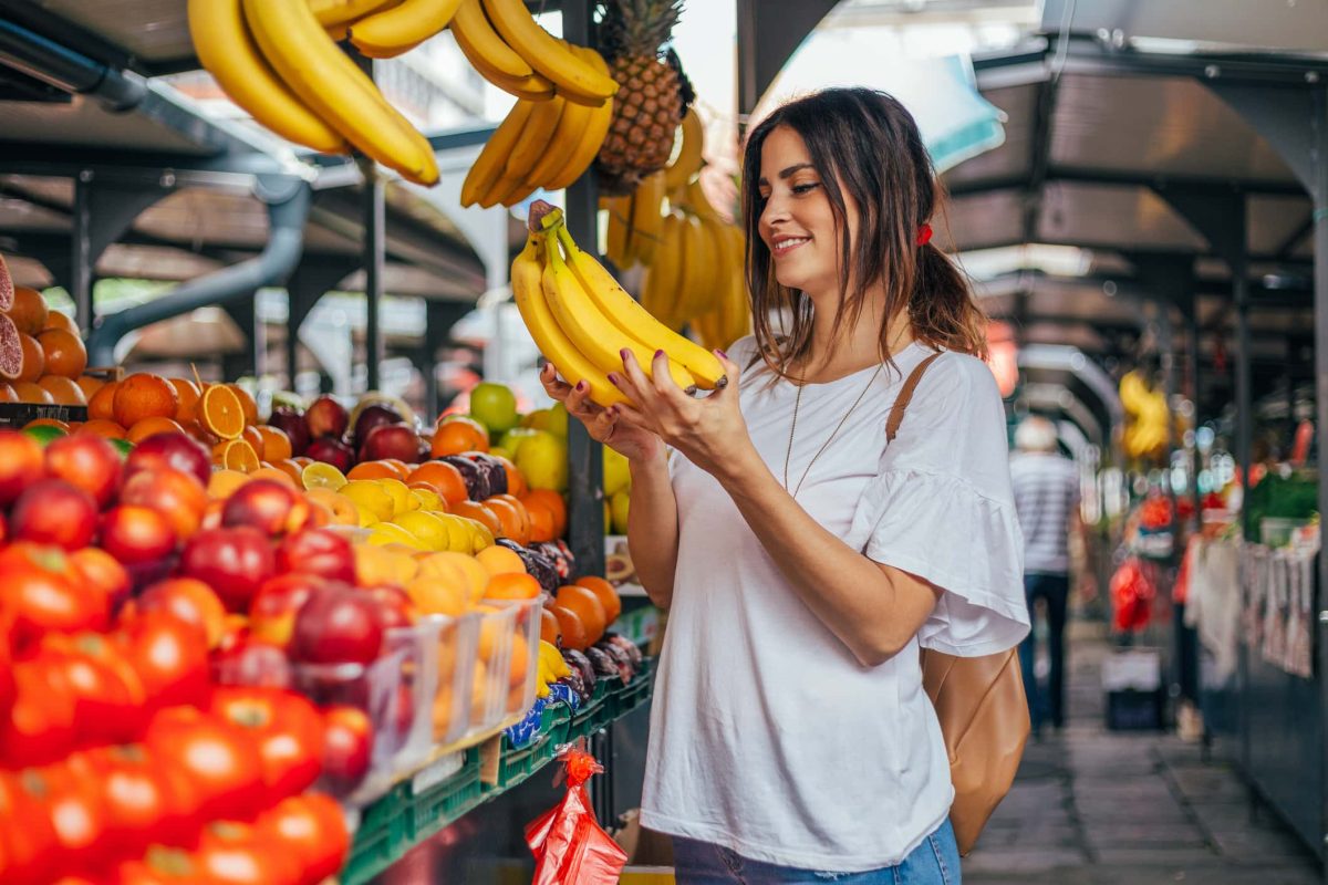 Eine Frau steht an einem Marktstand mit Obst und Gemüse und hält ein Bund Bananen in den Händen. Zu Hause hilft ein Tipp gegen braune Bananen: