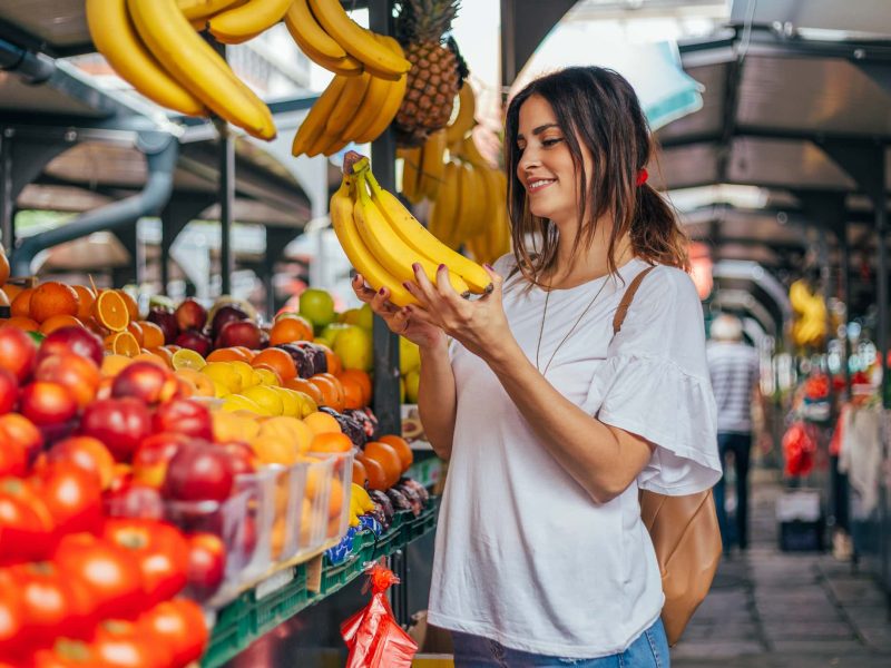 Eine Frau steht an einem Marktstand mit Obst und Gemüse und hält ein Bund Bananen in den Händen. Zu Hause hilft ein Tipp gegen braune Bananen: