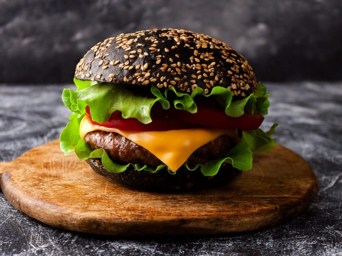Ein Burger mit Black Burger-Buns steht auf einem Holztab.lett auf einem Untergrund aus Stein. Im Hintergrund befindet sich eine Steinwand. Der Burger ist belegt mit Salat, Käse, Tomaten und einem Hackfleisch-Patty. Auf dem Burger-Bun befinden sich Sesamsamen.