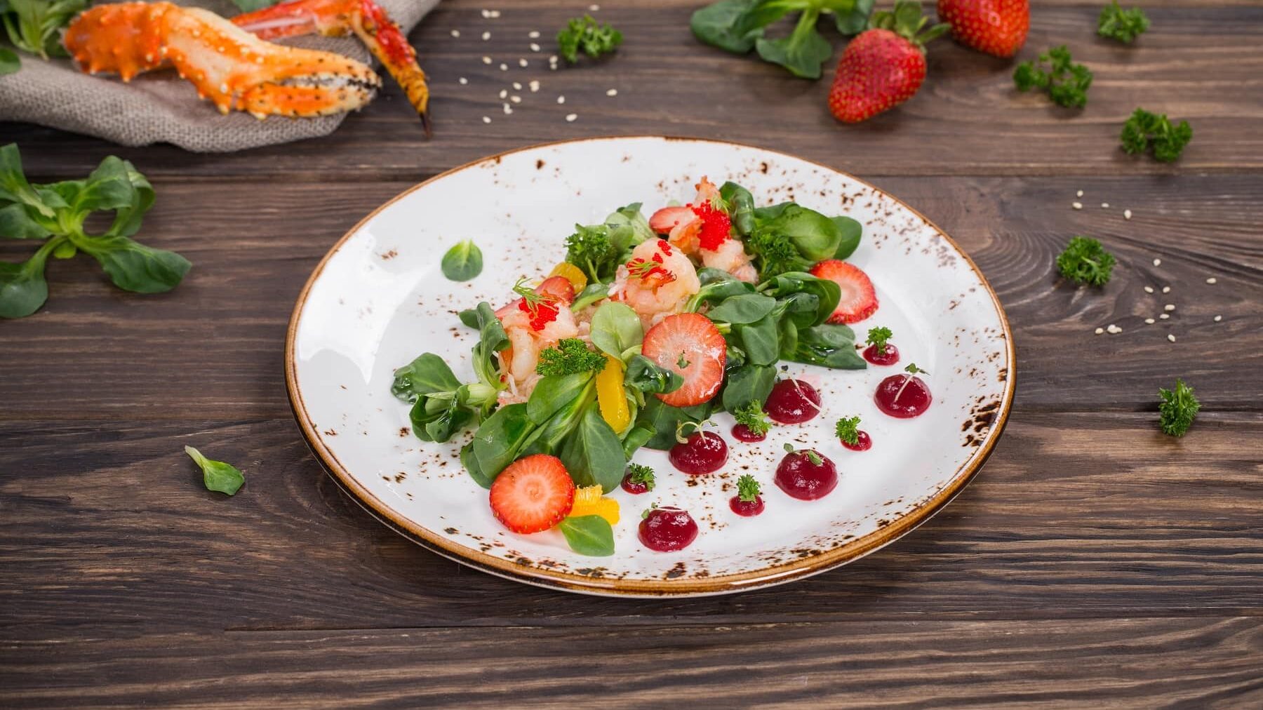 Ein weißer Teller mit Shrimps-Salatmit Erdbeeren steht auf einem braunen Tisch. Auf dem Tisch liegen noch Krabben, Salat, Petersilie und Erdbeeren.