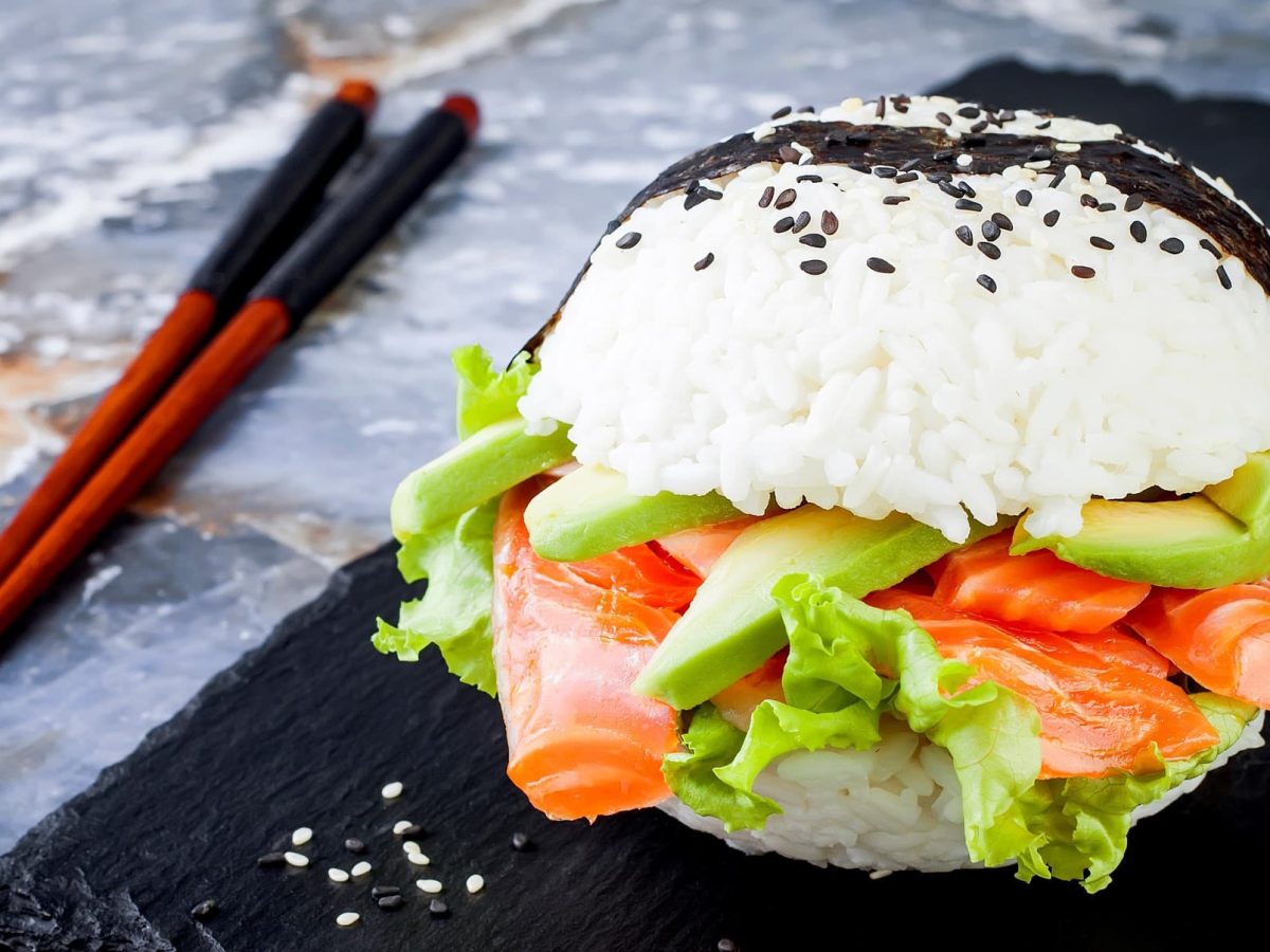 Ein Sushi-Burger mit Avocado und Lachs auf einer schwarzen Schiefernplatte. Daneben liegen Stäbchen.