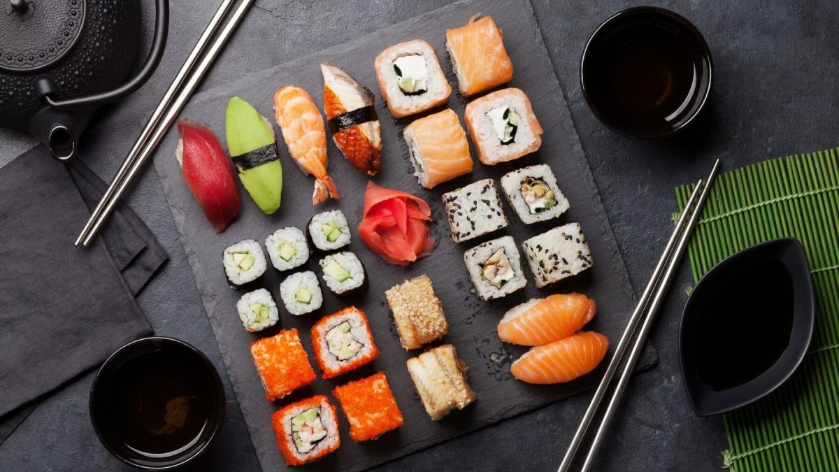 Verschiedenes Sushi liegt auf einer Steinplatte auf einem schwarzen Untergrund. Um die Platte herum stehen Schalen mit Sojasoße, eine Sushimatte und eine Teekanne. Neben dem Sushi liegen schwarze Essstäbchen.