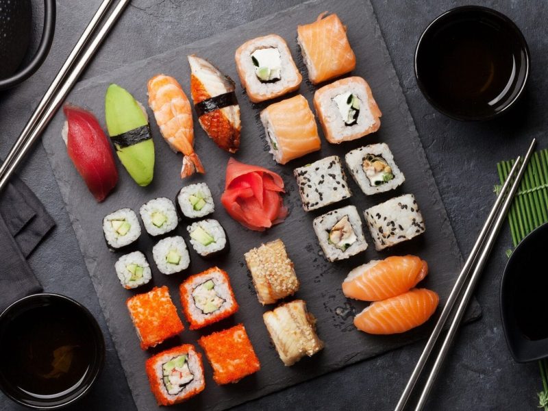 Verschiedenes Sushi liegt auf einer Steinplatte auf einem schwarzen Untergrund. Um die Platte herum stehen Schalen mit Sojasoße, eine Sushimatte und eine Teekanne. Neben dem Sushi liegen schwarze Essstäbchen.