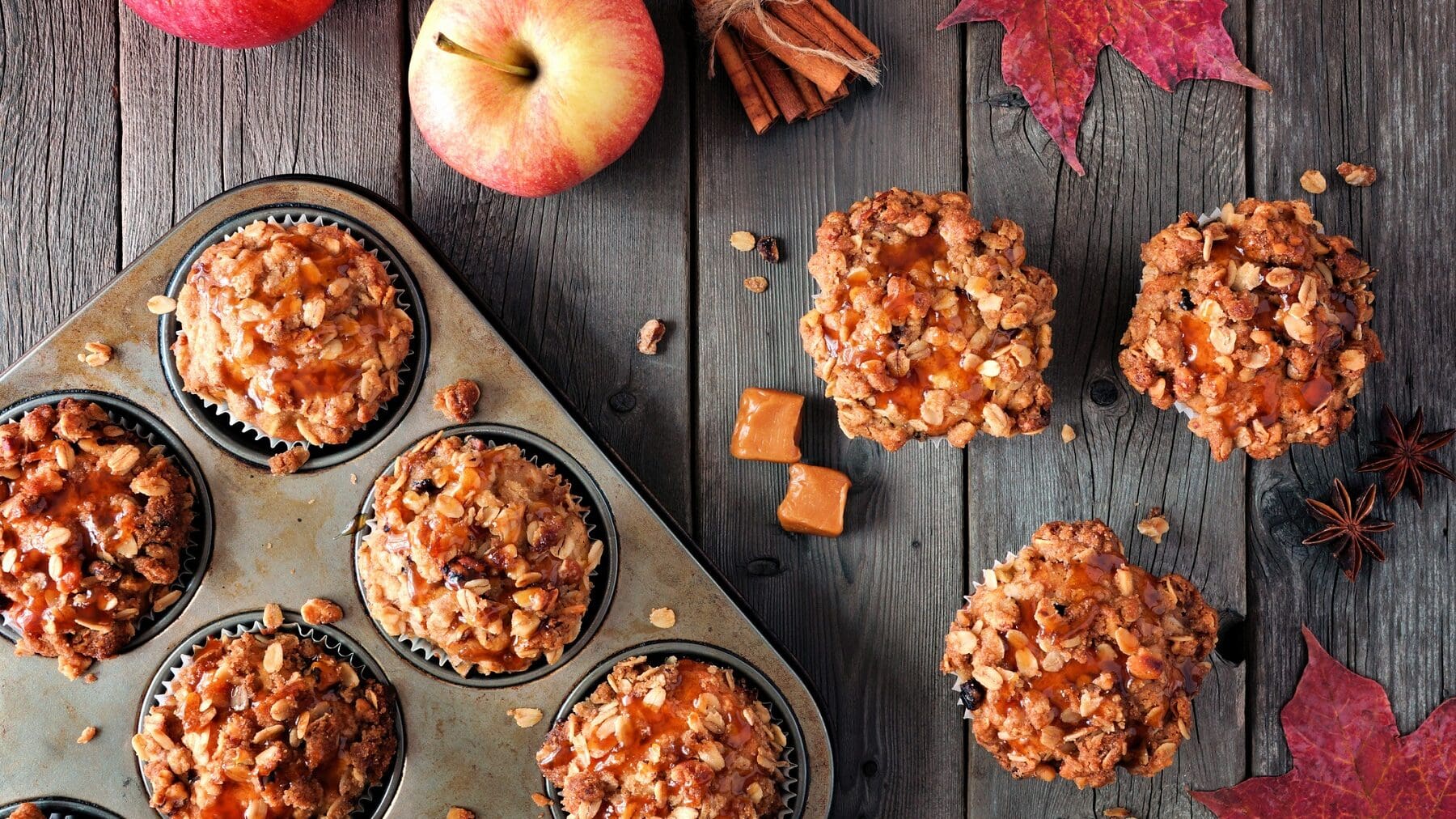 Drei Apfel-Karamell-Streusel-Muffins liegen auf einem grauen Holztisch. Neben ihnen steht eine Muffinform mit weiteren Muffins. Auf dem Tisch verteilt liegen Caramelwürfel, Haferflocken, Äpfel und Zimtstangen.