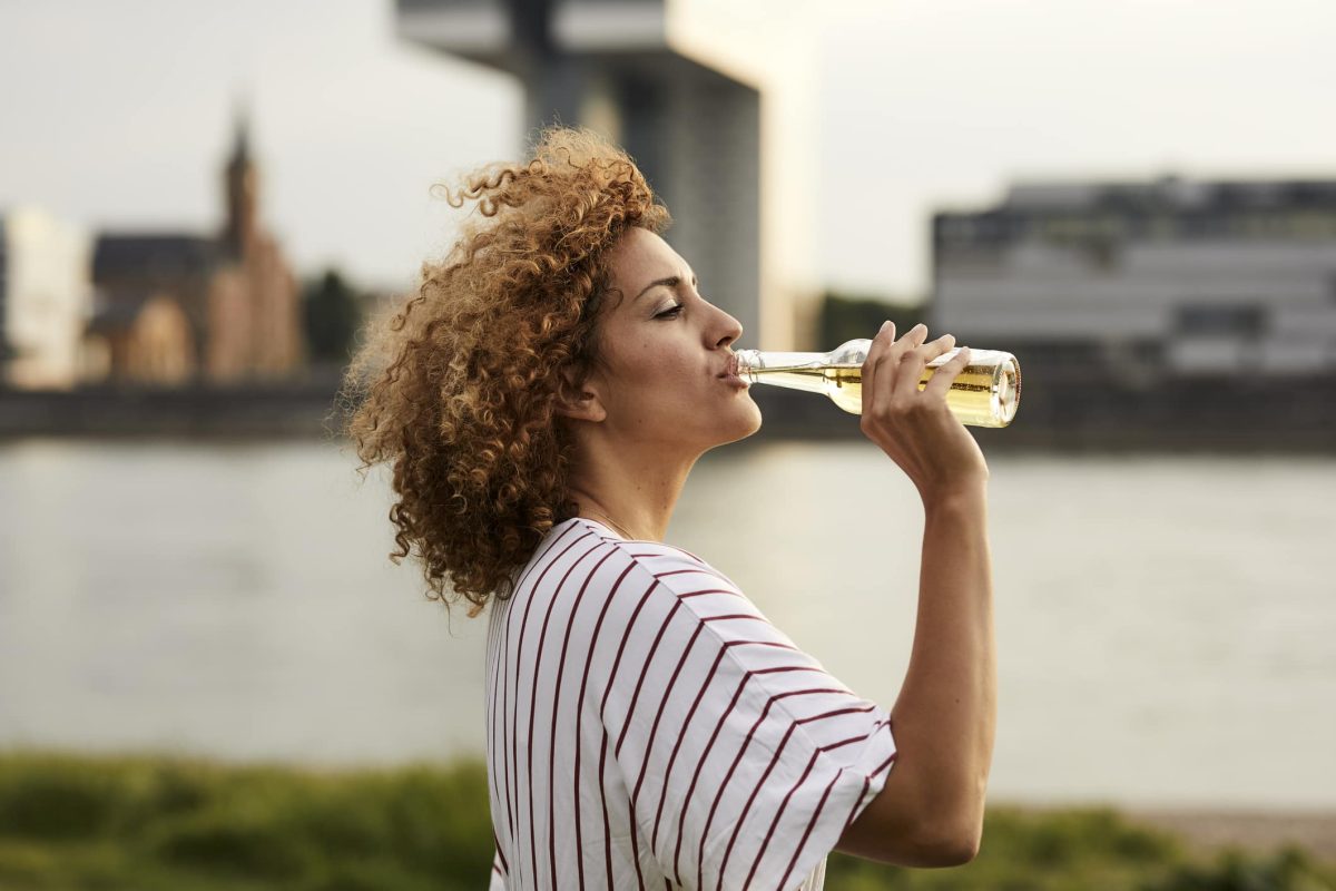 CacaoVida von Ritter Sport: Eine lockige Frau trinkt Limo aus einer Flasche, im Hintergrund ist unscharf ein Hafen zu erkennen.