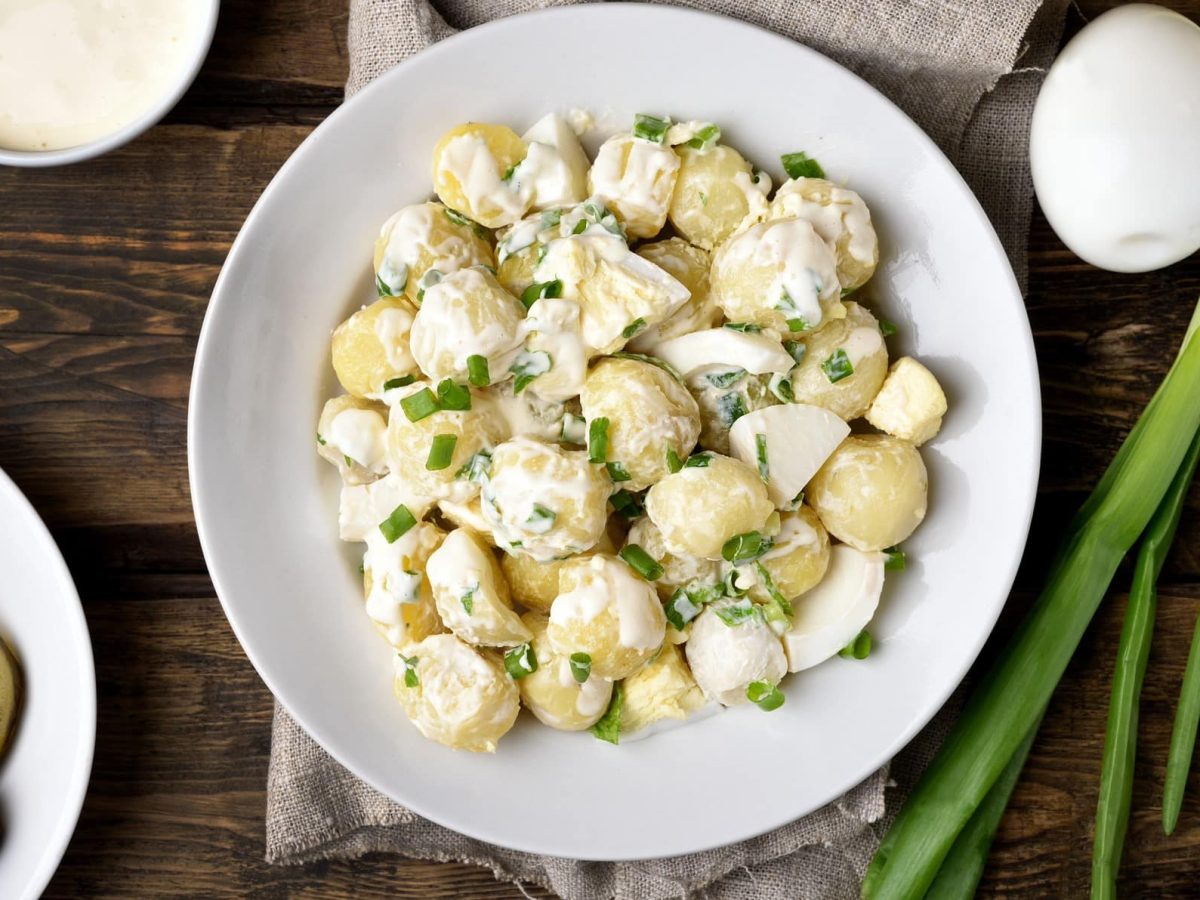 Kartoffelsalat mit Frühlingszwiebeln auf einem weißen Teller. Daneben liegen noch weitere Zutaten wie Eier, Frühlingszwiebeln und Gewürzgurken. Das Ganze auf einem hölzernen Untergrund.