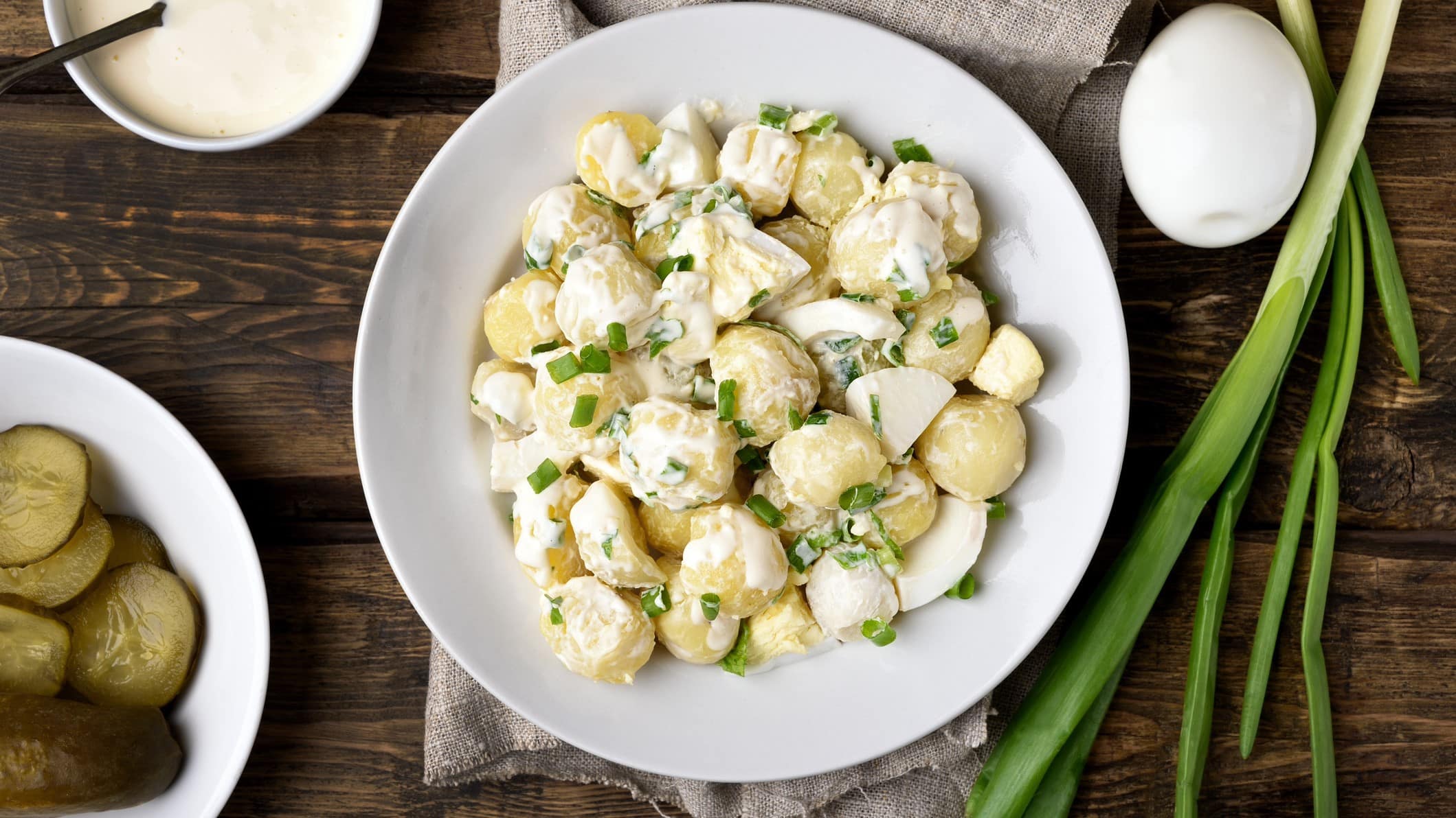 Kartoffelsalat mit Frühlingszwiebeln auf einem weißen Teller. Daneben liegen noch weitere Zutaten wie Eier, Frühlingszwiebeln und Gewürzgurken. Das Ganze auf einem hölzernen Untergrund.
