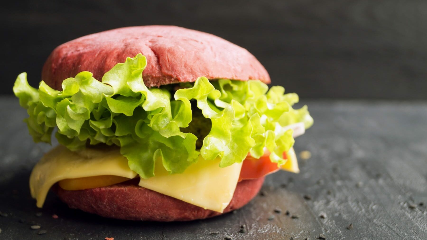 Ein Burger mit pinken Burger Buns, mit Salat, Käse und Tomaten steht auf einem schwarzen Holztisch. Auf dem Tisch verteilt liegen Sesamsamen. Der Hintergrund ist ebenfalls schwarz.