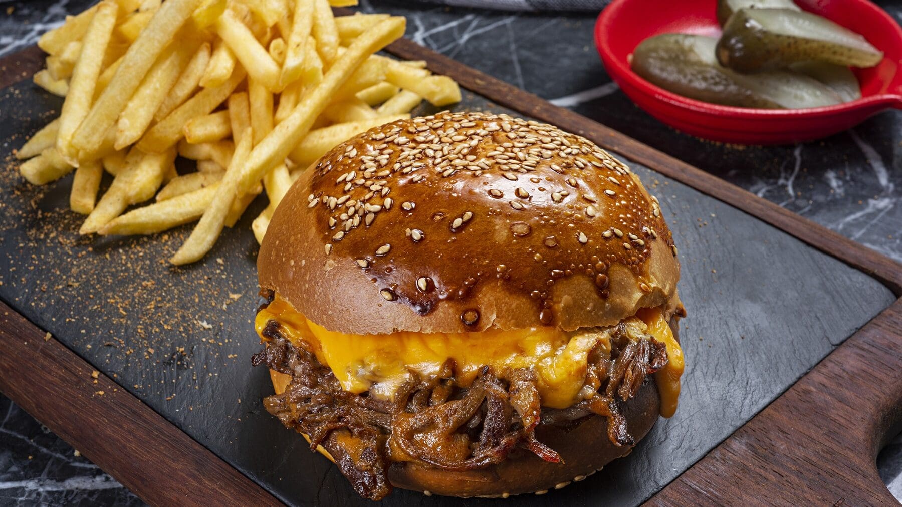 Ein Pulled Beef Burger mit Käsesauce steht auf einer Steinplatte auf einem schwarzen Tisch. Neben dem Burger liegen Pommes. Im Hintergrund steht eine rote Schale mit Gewürzgurken.