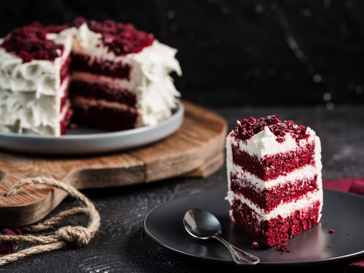 Ein Stück Red Velvet Cake ist auf einem schwarzen Teller angerichtet, zusammen mit einem Löffel. Im Hintergrund sieht man den ganzen Kuchen, dem ein Stück fehlt, auf einem Holzbrett stehen.