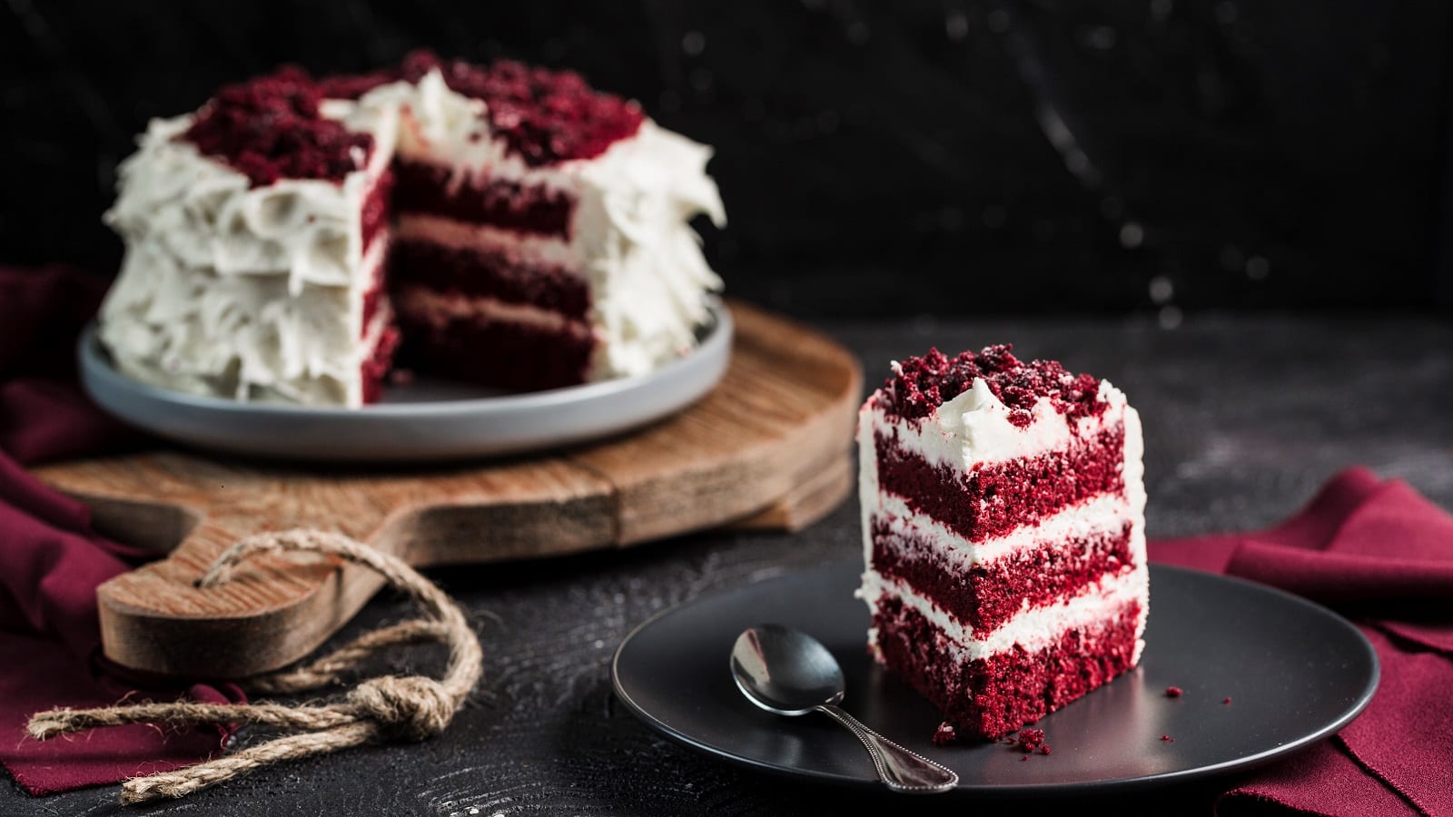 Ein Stück Red Velvet Cake ist auf einem schwarzen Teller angerichtet, zusammen mit einem Löffel. Im Hintergrund sieht man den ganzen Kuchen, dem ein Stück fehlt, auf einem Holzbrett stehen.