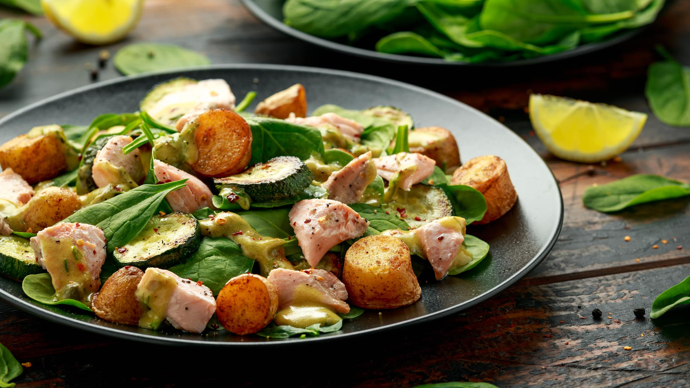 Ein Teller Salat mit Lachs, Blattspinat und Kartoffeln, daneben liegen Spinatblätter und Zitronenscheiben.