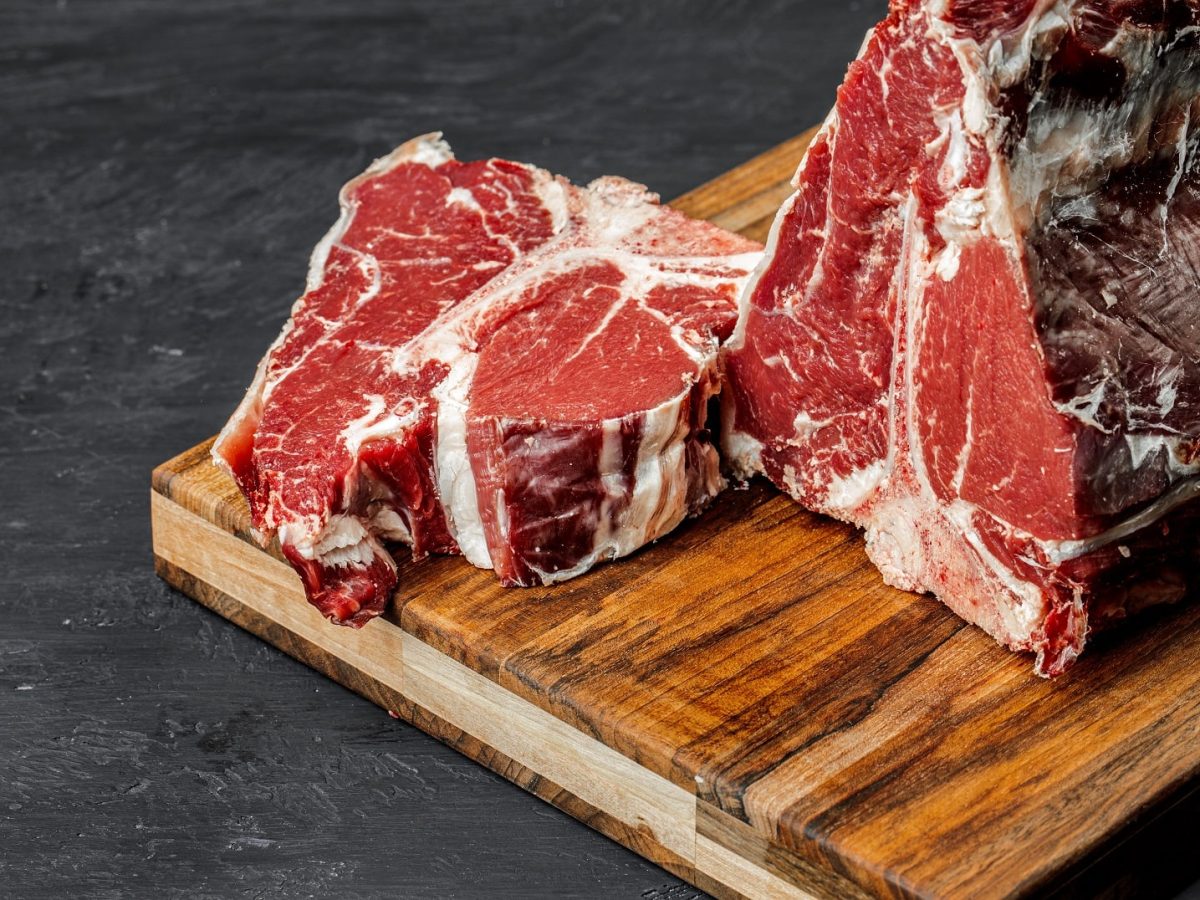 Ein rohes Stück T-Bone-Steak wurde von einem größeren Fleischstück abgeschnitten und liegt auf einem Holzbrett.
