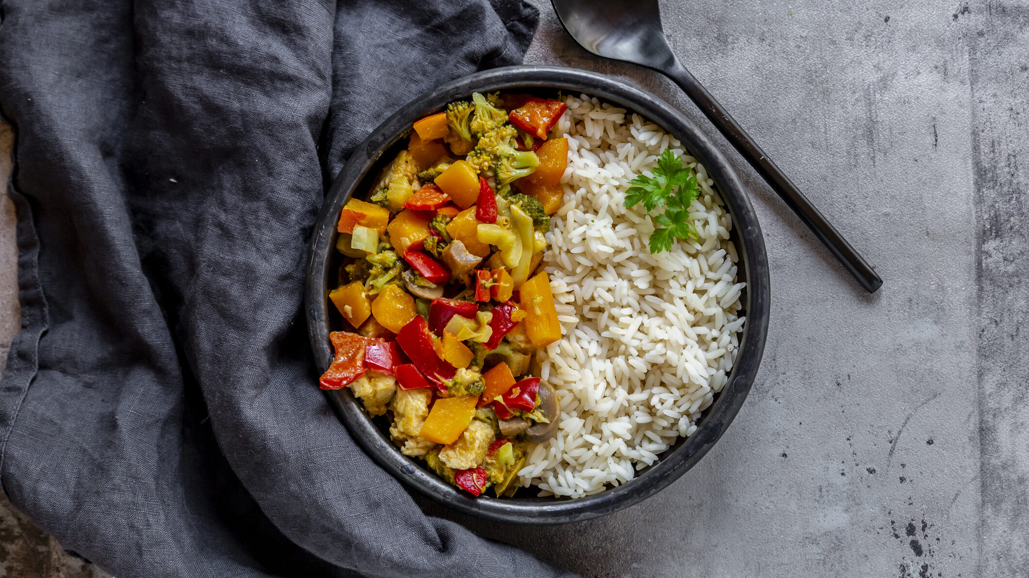 Eine dunkle Schale mit vegetarischem Gemüsecurry und Reis in der Draufsicht, daneben liegen ein schwarzer Löffel und ein graues Geschirrtuch.