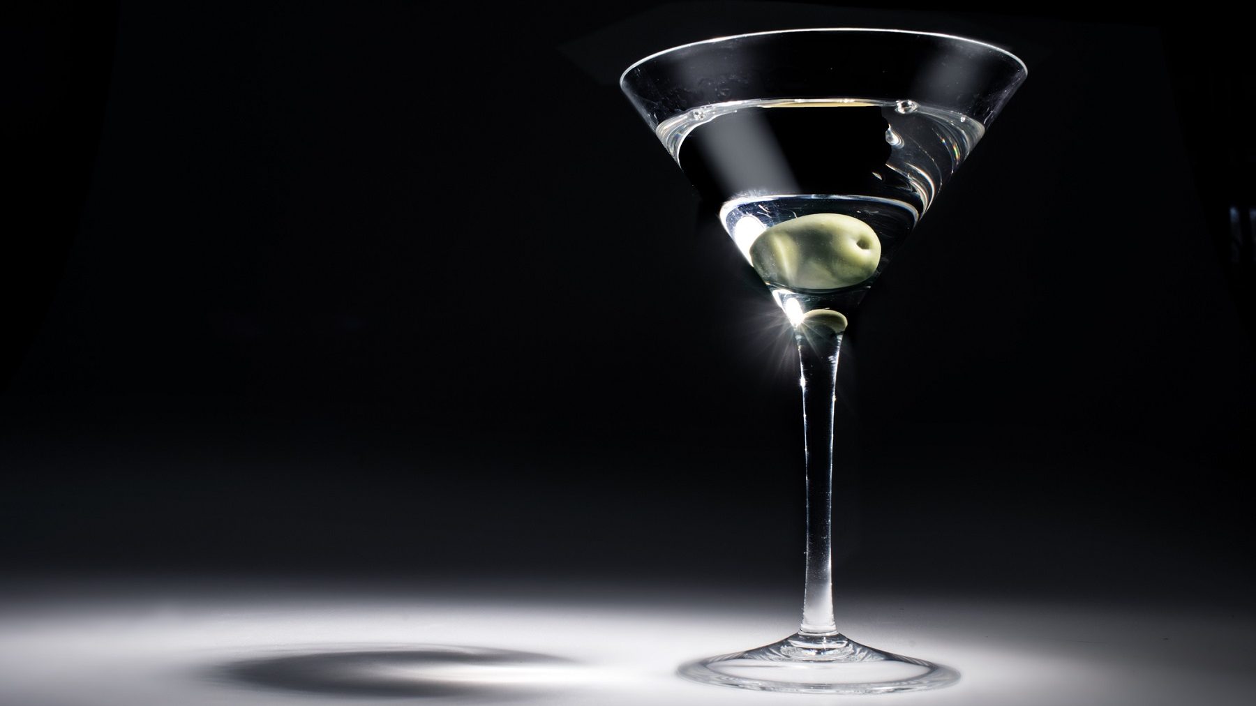 James Bonds Martini mit einer Olive in einem Martiniglas. Das Glas wird aus einer Richtung stark beleuchtet, der Hintergrund und Boden sind dunkel.