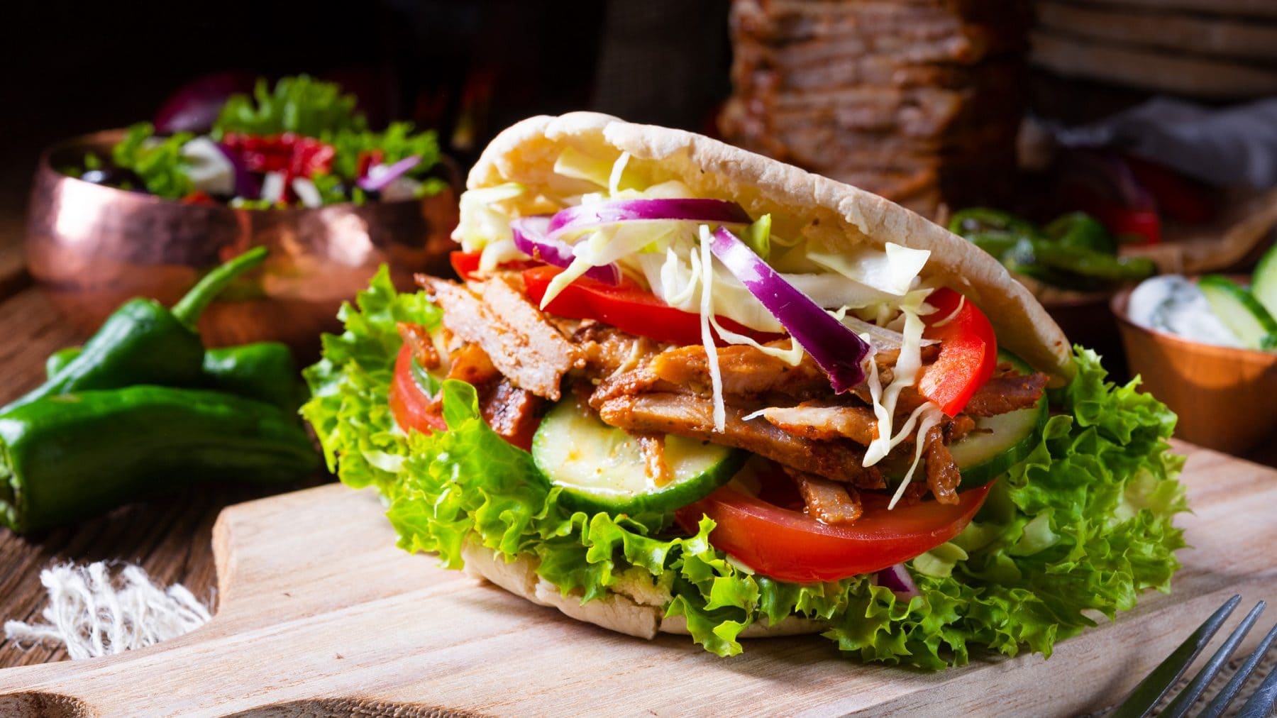 Eine Portion Avengers Shawarma liegt auf einem Holzbrett. Im Hintergrund ist ein Spieß mit Fleisch, Paprika, Salat und einige Gurkenscheiben,