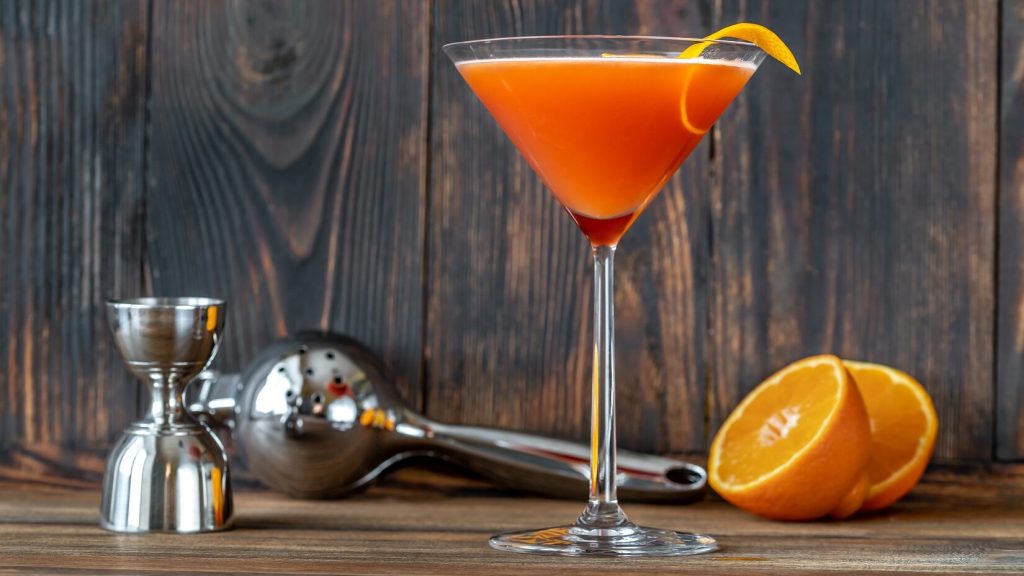 Der Blood and Sand-Cocktail eingegossen in einem Martini-Glas. Im Hintergrund liegt eine aufgeschnitte Orange sowie eine Presse und ein Messbecher.