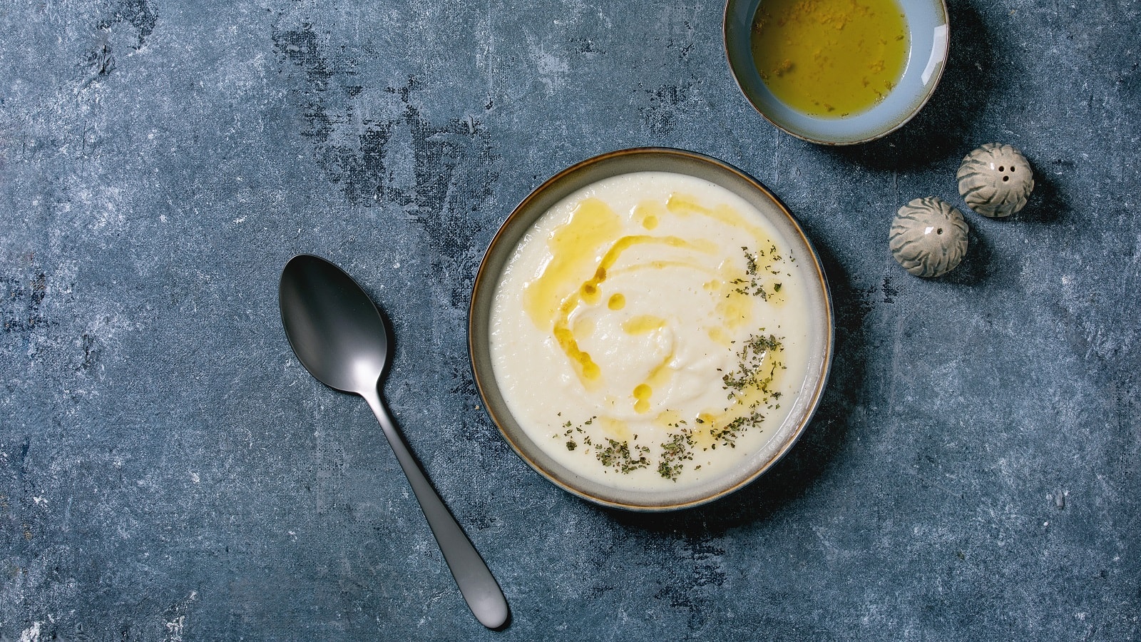 Eine Schüssel mit einer Portion weißer Fenchel-Creme-Suppe garniert mit Gewürzen und Olivenöl neben salz und Pfefferstreuer, einer kleinen Schüssel Olivenöl sowie einem Löffel, vor grauem Hintergrund von oben.