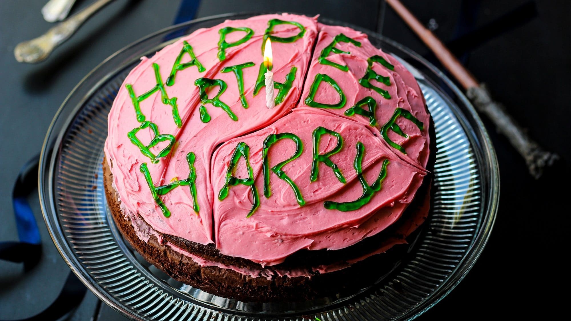 Harry Potters Geburtstagskuchen mit Schokoteig, pinker Buttercreme und grüner Zuckerschrift auf einem Glasteller auf einem schwarzen Tisch. Im Hintergrund liegt ein Zauberstab.