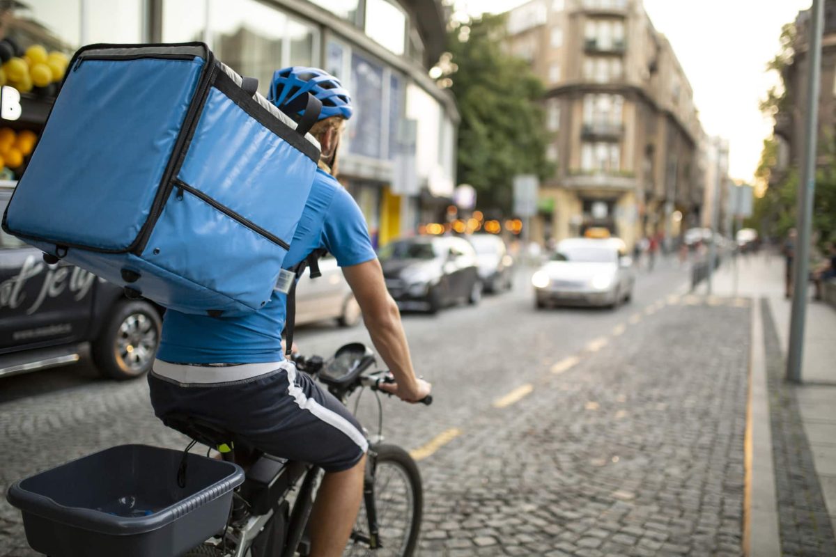 Wuplo: Ein Fahrradkurier, der von hinten zu sehen ist und einen großen Rucksack trägt, auf einer gepflasterten Straße.