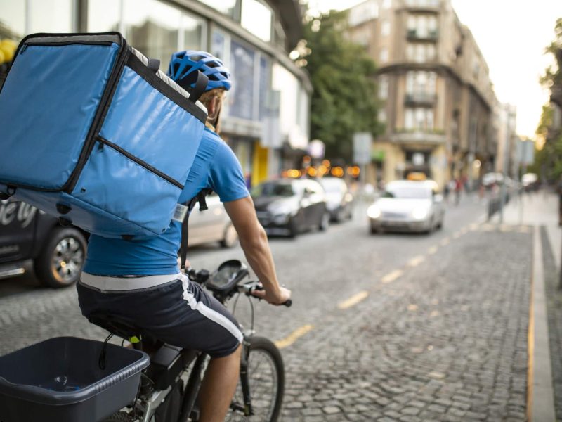 Wuplo: Ein Fahrradkurier, der von hinten zu sehen ist und einen großen Rucksack trägt, auf einer gepflasterten Straße.