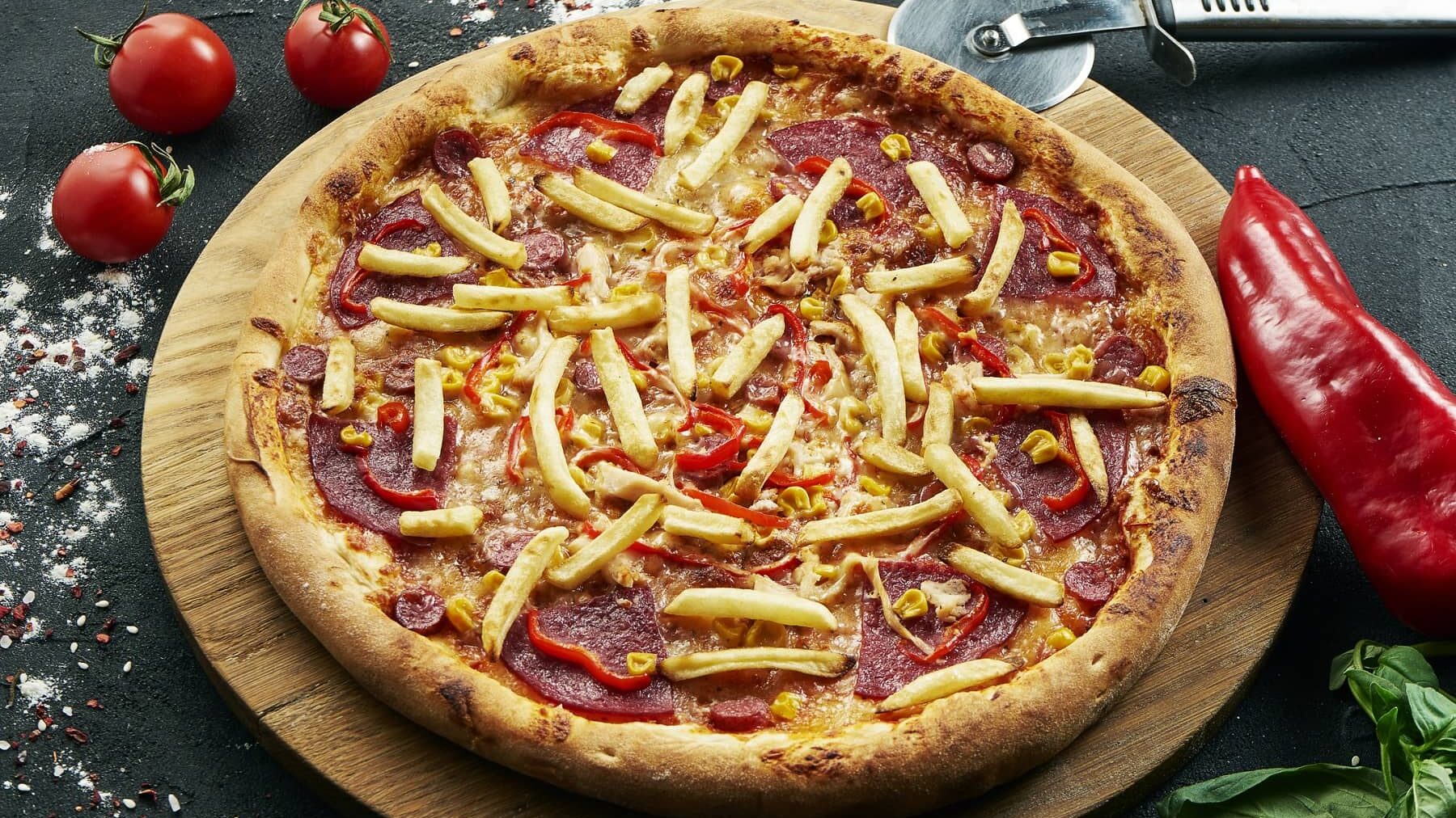 Eine Pommes-Pizza mit Salami-Paprika und Mais auf einem runden Holzbrett auf einem Steintisch. Um die Pizza herum liegen Paprika, Tomaten, ein Pizzaroller und Basilikum.