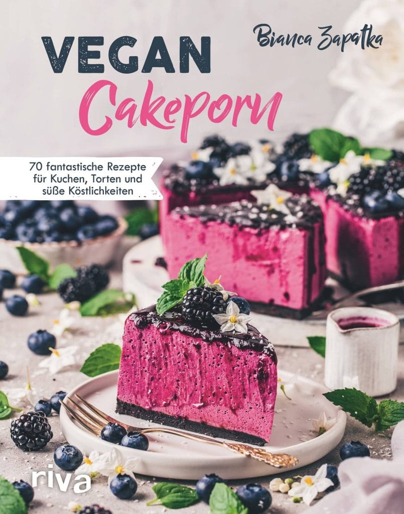 Das Buchcover von Vegan Cakeporn von Bianca Zapatka, auf dem ein Stück Blaubeer-Käsekuchen zu sehen ist. Im Hintergrund sieht man den restlichen Kuchen sowie frische Beeren, die auf dem Tisch liegen.