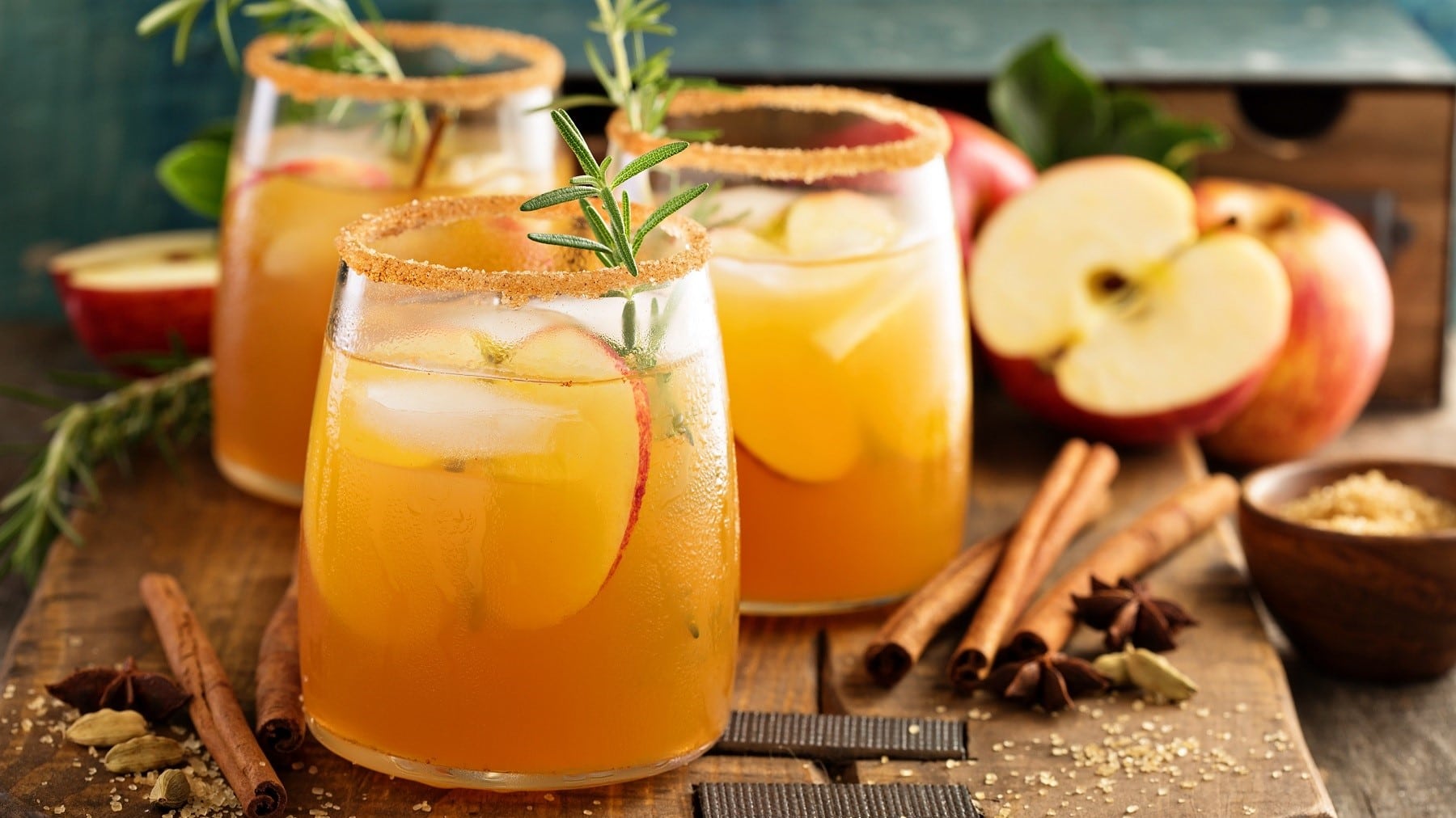 Drei Gläser Apple-Cider-Cocktail mit Zimt und Zucker am Rand. Die Cocktails sind angerichtet mit Eis, Apfel und Gewürzen. Umdie Gläser herum liegen herbstliche Gewürze, Zimtstangen, Zimt und Zucker. Im Hintergrund liegen Äpfel. Die Gläser stehen auf einem braunen Holzbrett.