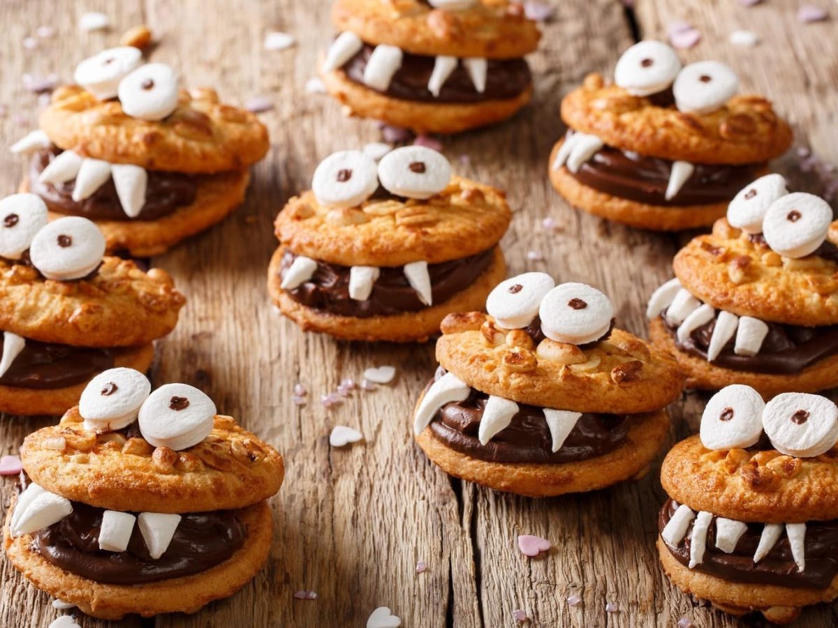 Mehrere Monster-kekse mit Marshmallow-Augen und Schokocreme liegen auf einem Holztisch. Um die Monster herum liegen Streusel in Herzform.