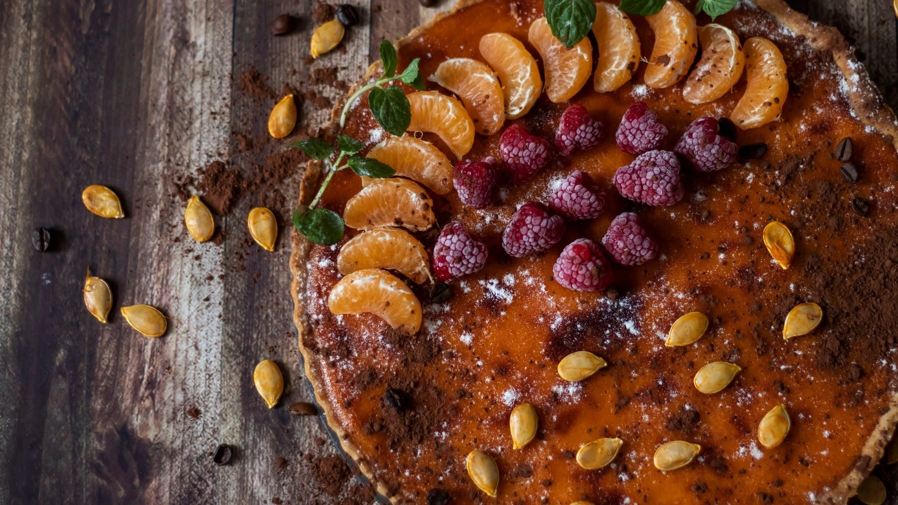 Ein Pumpkin Pie mit Obst ist angerichtet mit Mandarinen, Himbeeren, Kakaopulber und Puderzucker. Auf dem Kuchen und dem Untergrund liegen Kürbiskerne verteilt. Der Untergrund beseht aus braunem Holz.