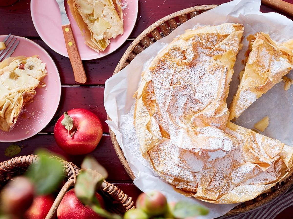 Ein angeschnittener Apfelstrudel-Kuchen auf einem weißen Backpapier neben zwei kleinen rosa Tellern mit jeweils einem Stück des Kuchens und einer Gabel, und frischen Äpfeln und einem Küchentuch, vor einem dunkel roten Holzhintergrund, von oben.