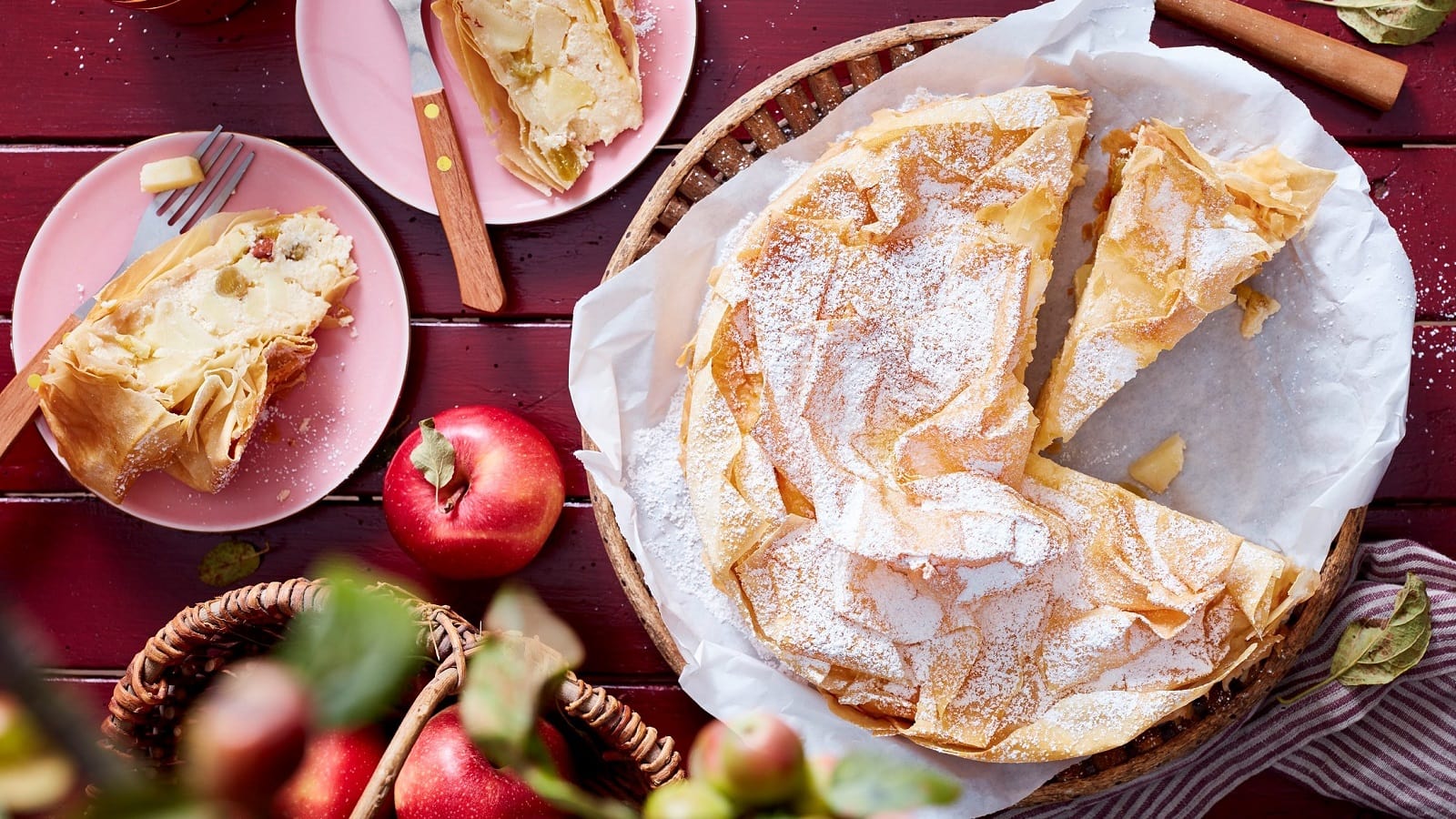 Ein angeschnittener Apfelstrudel-Kuchen auf einem weißen Backpapier neben zwei kleinen rosa Tellern mit jeweils einem Stück des Kuchens und einer Gabel, und frischen Äpfeln und einem Küchentuch, vor einem dunkel roten Holzhintergrund, von oben.