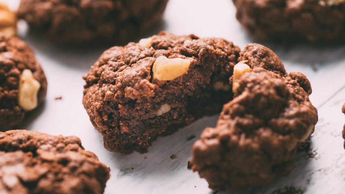Ein angeschnittener Chocolate Chip Cookie mit dreierlei Schokolade auf hellem Holzuntergrund, drumherum sieht man unscharf ein paar weitere Kekse.