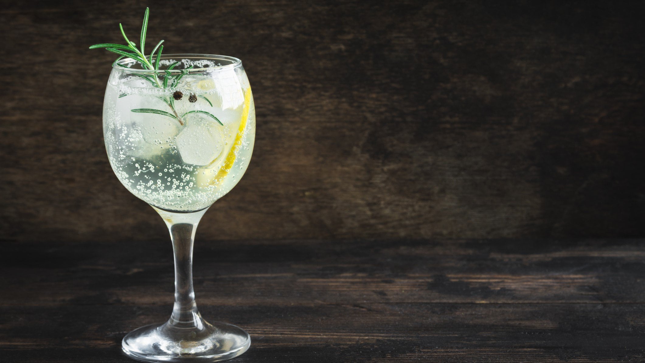 Ein Glas Gin Tonic Spritz mit einer Zitronenscheibe, Rosmarin und Eiswürfeln, vor einem dunklen Hintergrund.