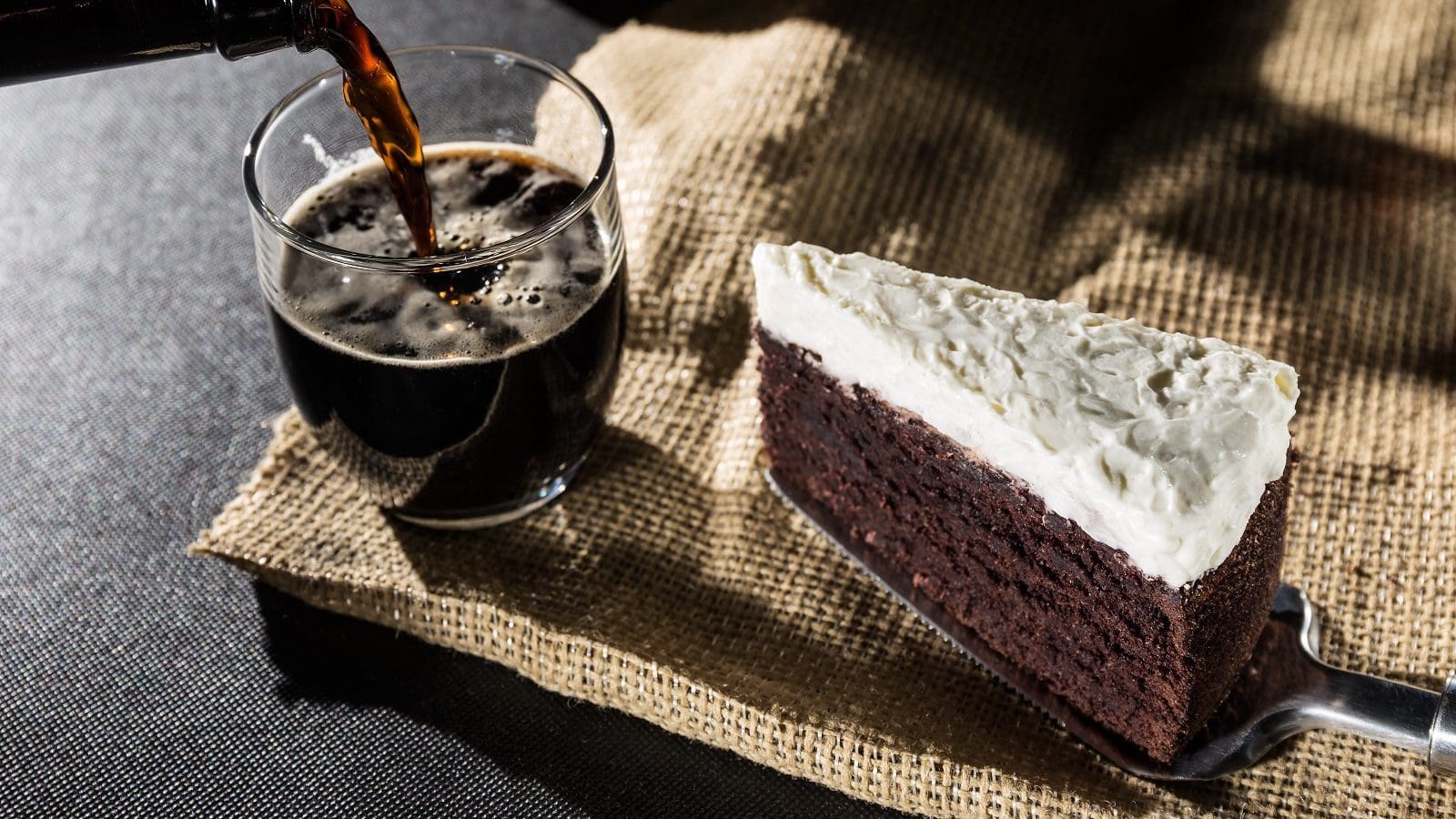 Ein Stück Guinness-Cake auf einem Kuchenheber neben einem Glas, in das gerade Guinness eingegossen wird, beides auf einem groben, braunen Tuch vor einem dunklen Hintergrund.