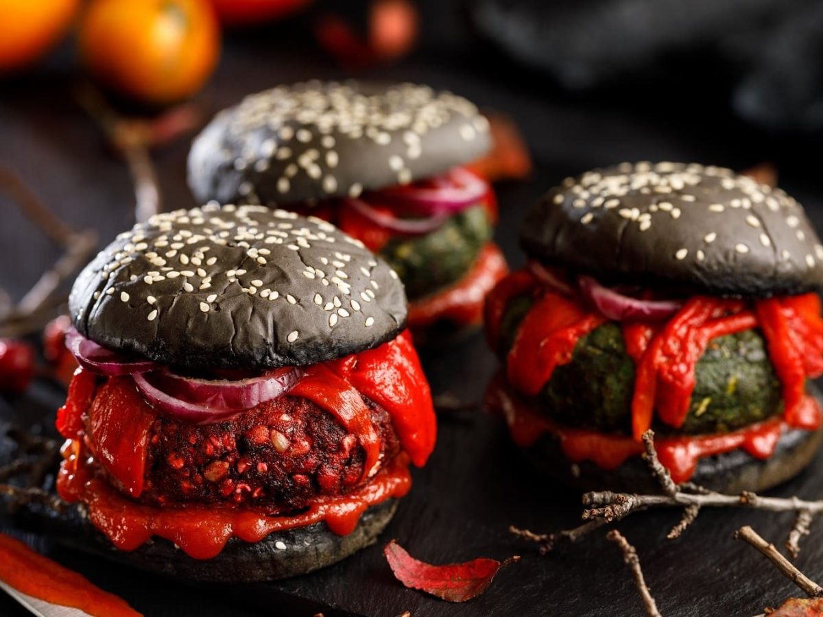 Drei Halloween-Burger mit verschiedenen Patties stehen auf einem Untergrund aus schwarzem Holz. Um die Burger herum liegen herbstliche Blätter und trockene Äste.