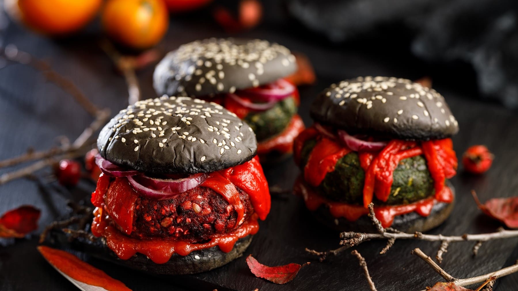 Drei Halloween-Burger mit verschiedenen Patties stehen auf einem Untergrund aus schwarzem Holz. Um die Burger herum liegen herbstliche Blätter und trockene Äste.