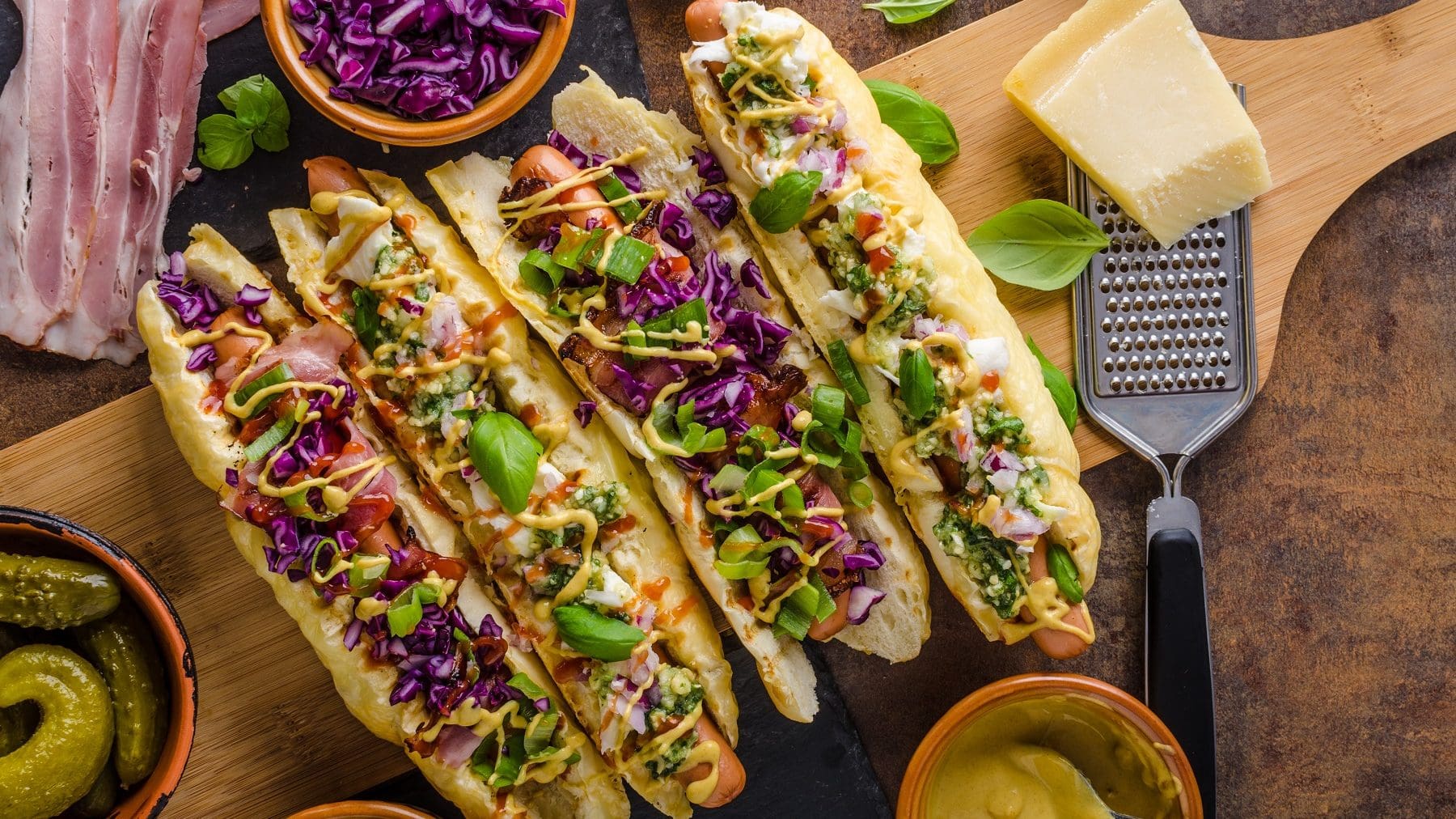 Hot Dogs mit Gemüse und Curry-Sauce auf einem Holzbrett. Die Hot Dog Brötchen sind mit Käse überbacken. Um die Hot Dogs herum liegt Bacon, vier Schalen mit Gurken, Lauch, Rotkohl und Curry-Sauce. Rechts neben den Brötchen liegt ein Stück Käse mit einem Käsehobel. Über den Hot Dogs ist Basilikum verteilt.