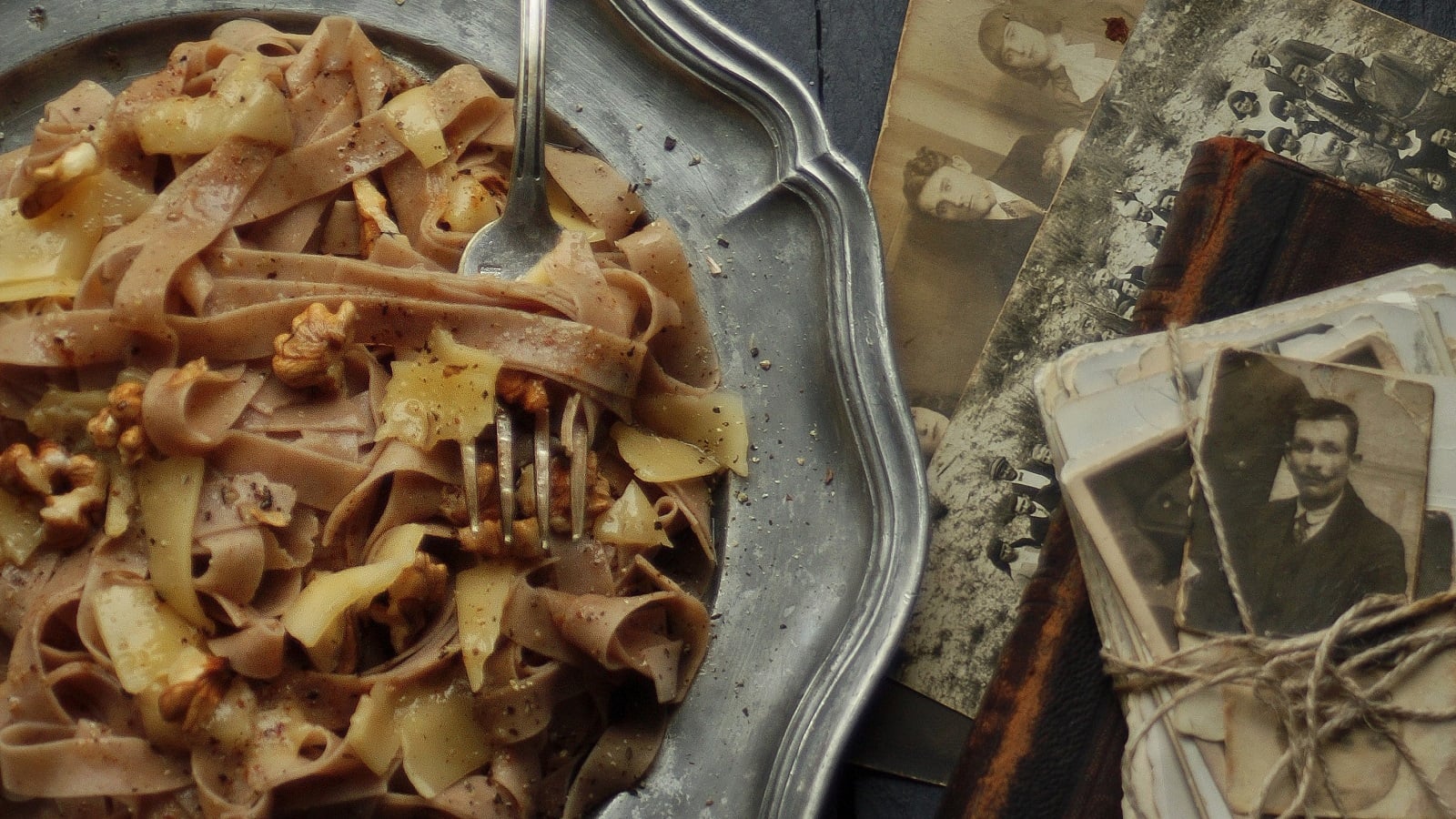Ein silbener Teller mit einer Portion selbstgemachter Kastanien-Pasta mit Walnusssoße, garniert mit Parmesanscheiben und Walnüssen neben alten Fotografien und Büchern vor einem dunklen Hintergrund, von oben.