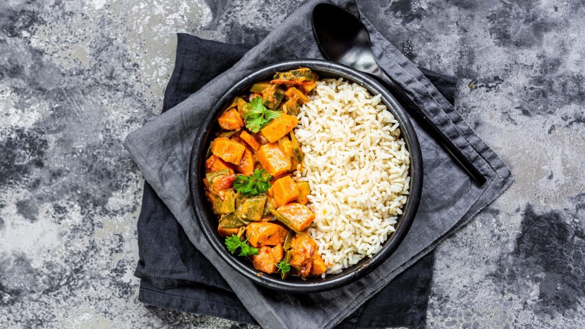 Ein dunkler Teller Reis und Kürbis-Curry mit Lauch steht auf zwei grauen Geschirrtüchern auf einem Marmortisch, Draufsicht.