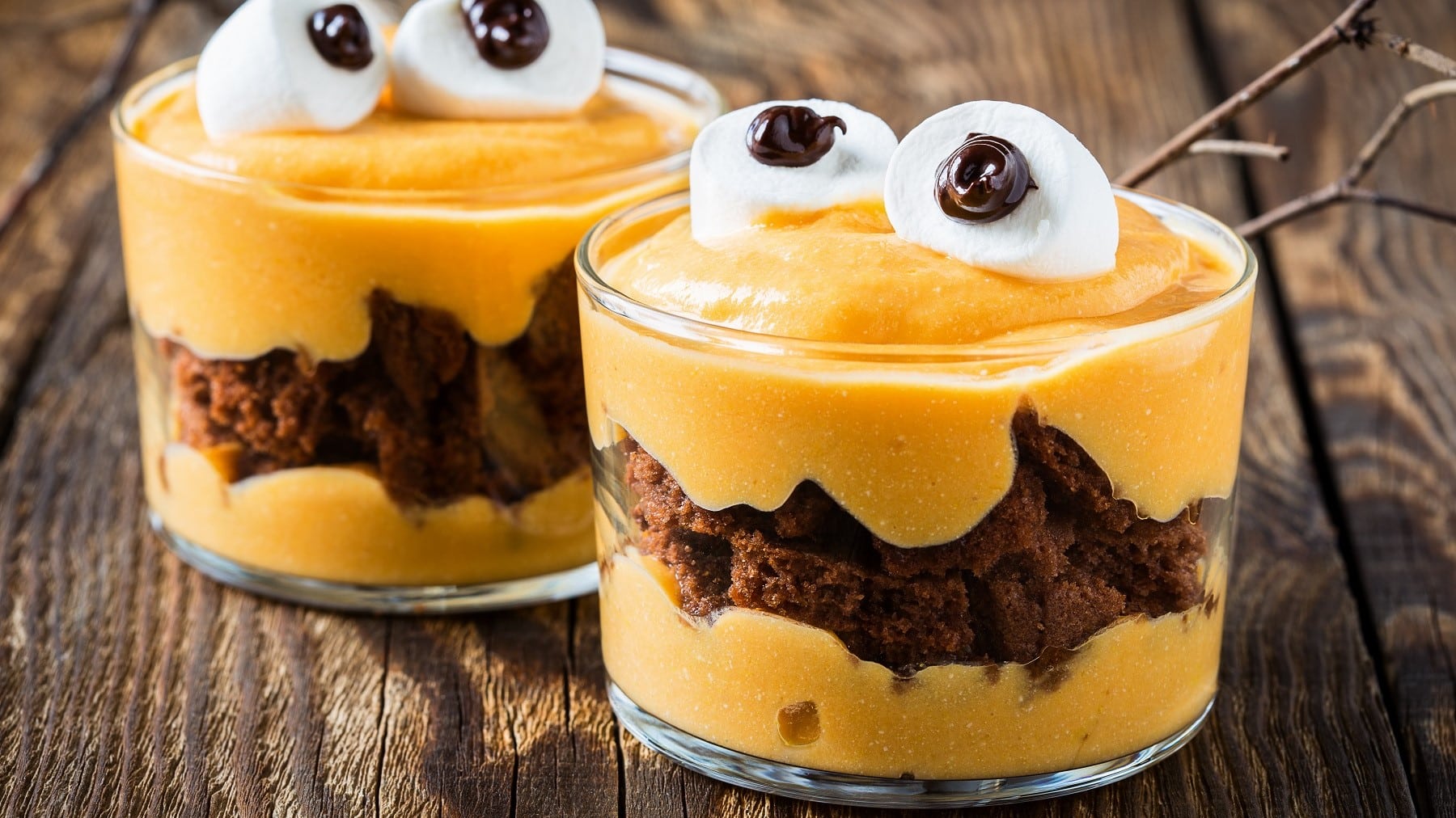 Zwei Gläser mit Monster-Dessert stehen auf einem braunen Holztisch. Das Dessert besteht aus Schokokuchen, Kürbispüree und Marshmallows als Augen. Hinter den Gläsern liegt ein Ast als Dekoration.