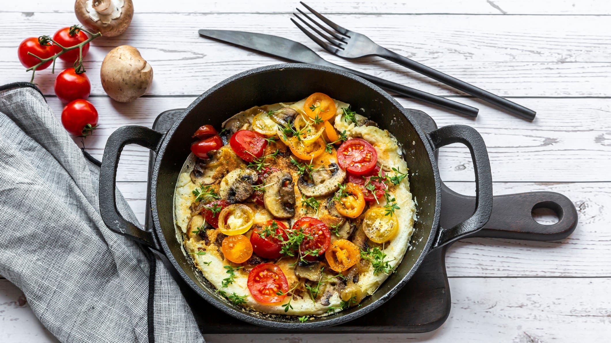 Eine gusseiserne Pfanne mit einem Omelett mit Pilzen und Tomaten sowie Oregano in der Draufsicht auf hellem Untergrund. Daneben liegen Besteck, ein paar Tomaten und Pilze sowie ein Geschirrtuch.