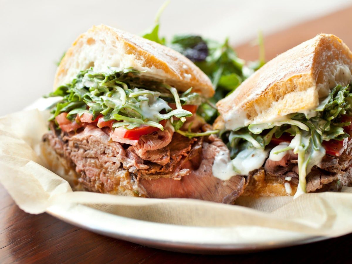 Ein halbiertes Roastbeef-Sandwich mit Rucola auf einem Teller, der auf einem Holztisch steht.