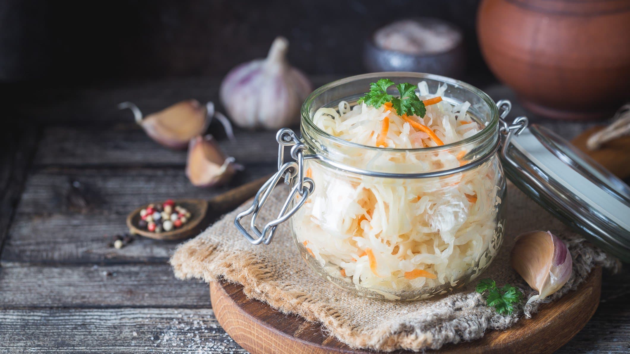 Ein Einmachglas mit Sauerkrautsalat mit Möhren, das auf einem runden Holzbrett steht. Drumherum sieht man Zutaten wie Knoblauch und Pfefferkörner.