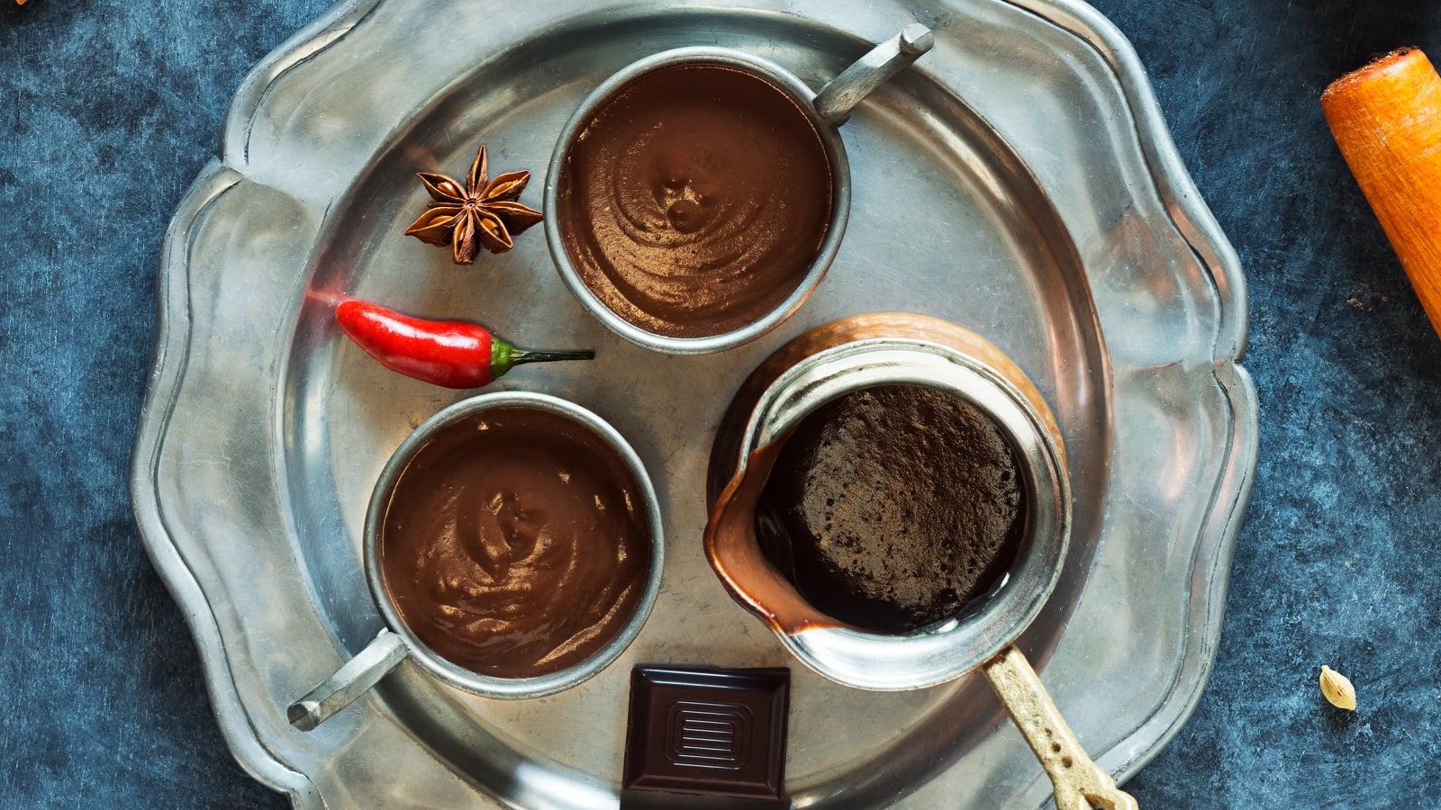 Zwei Tassen gefüllt mit Xocolatl neben einer Kanne, einem Stück zartbitterschokolade, einer Chilischote und einem Stück Sternanis auf einem silbernen Tablett vor einem dunkelblauem Hintergrund, von oben.