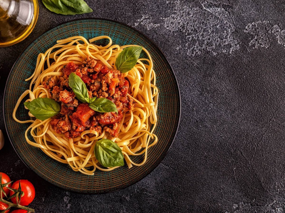 Teller Spaghetti Bolognese umgeben von verschiedenen Soßen-Zutaten auf dunklem Untergrund.