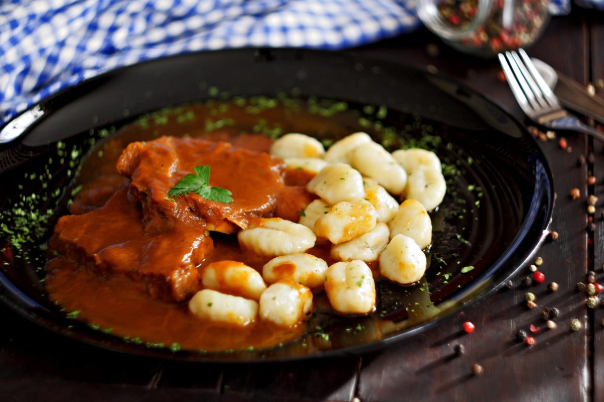 Der Dalmatinische Rinderbraten angerichtet mit Kartoffelgnocchi auf einem schwarzen Teller. Dazu Pfefferkörner als Deko.