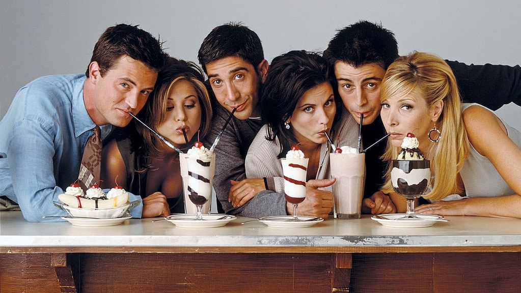 Charaktere aus der Serie Friends zusammen in der Frontalansicht am Tisch. Alle Gesichter dem Betrachter zugewandt. Je drei trinken gemeinsam aus einem Glas mit Milkshakes. Auf dem Tisch drei Milchshakes, ein Bananasplit-Eis und ein Eisbecher mit Schoko- und Vanilleeis.